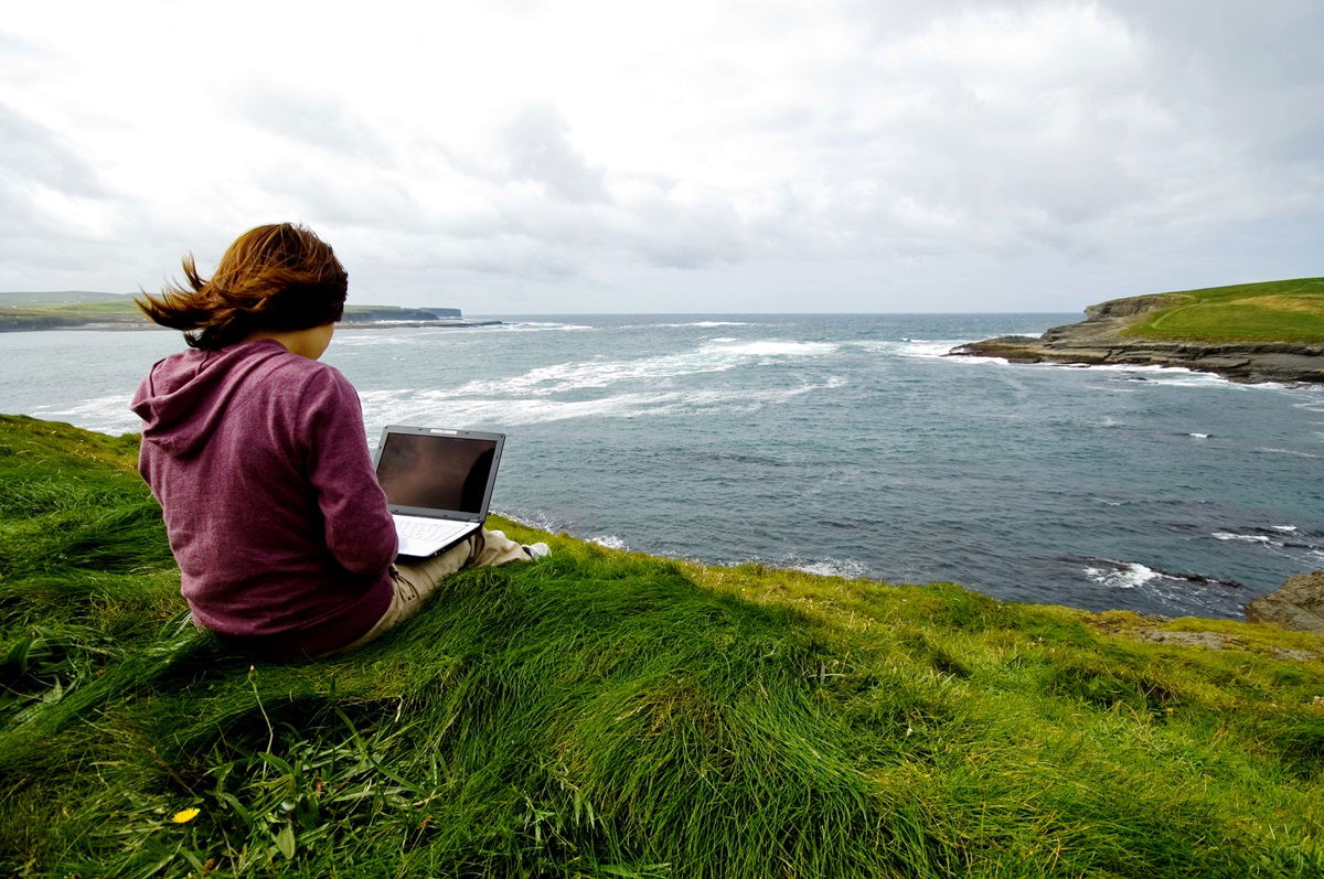 Frau sitzt auf einem grasbedeckten Berg mit Laptop am Meer