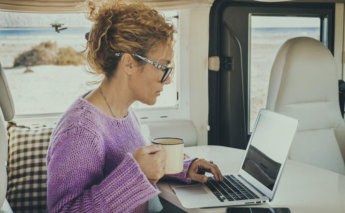 Frau arbeitet am Laptop im Camper mit Strand und Meer im Hintergrund