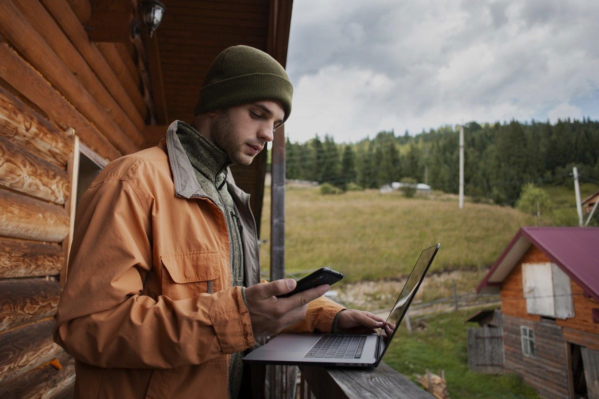 Mann sitzt mit Laptop vor einer Blockhütte in den Wäldern von Kanada