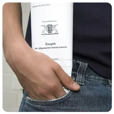 Detail view of a right trouser pocket in which one hand is inserting a piece of paper marked "Zeugnis der allgemeinen Hochschulreife".