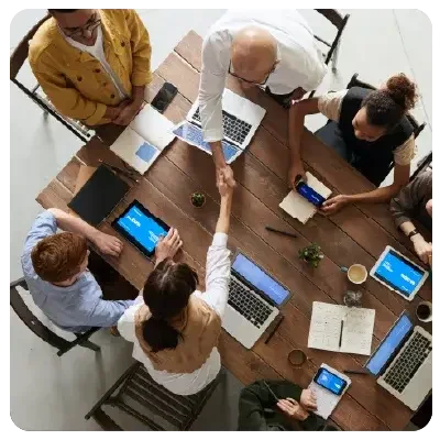 A total of 6 men and women sit at a brown wooden table with laptops, smartphones, tablets and notebooks on the table in front of them. A man and a woman shake hands across the table. 