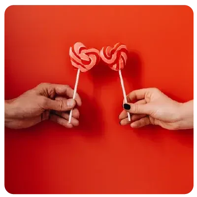 Vor einem roten HIntergrund sind zwei Hände - jeweils einer Frau und eines Mannes - die beide ein Zuckerstangenherz aneinander halten.