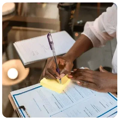 An einem Glastisch sitzend notiert eine Frau in rosa Bluse etwas auf einem gelben Post-it Zettel, der auf einem tabellarischen Lebenslauf liegt. Daneben liegt ein beschriebenes Notizbuch.