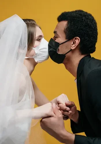 Eine Braut mit weißem Schleier und weißer Mund-Nasen-Maske "küsst" ihren Bräutigam, der ganz in schwarz gekleidet ist und eine schwarze Maske trägt.