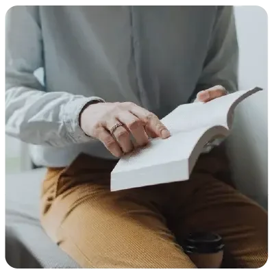 Un hombre con pantalones marrones y camisa clara se sienta con un libro abierto en la mano y señala dentro del libro.