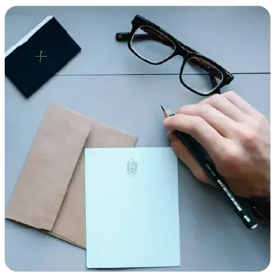A brown envelope, a pair of glasses and blank stationery lie on a white background. To the right is a right hand holding a pen.