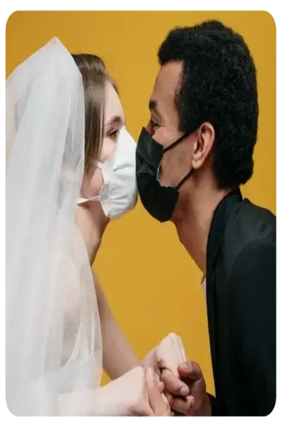 A bride with white veil and white mouth-nose mask "kisses" her groom, who is dressed all in black and wears a black mask.