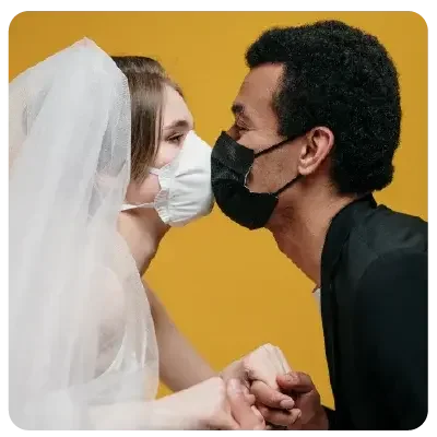 A bride with white veil and white mouth-nose mask "kisses" her groom, who is dressed all in black and wears a black mask.