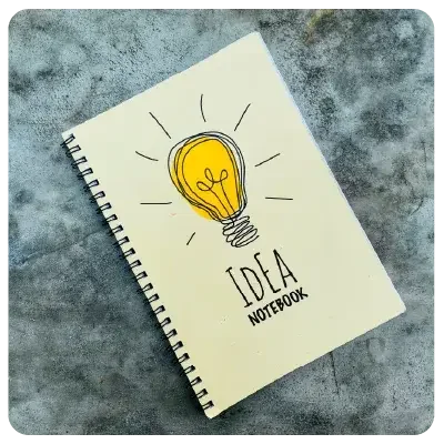 Kladde lies on a grey patterned background. On the cover is a bright light bulb drawing and the text "IDEA Notebook".