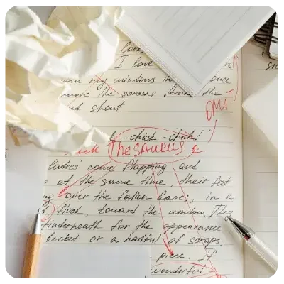 Vista de un papel completamente escrito en el que se aprecian las correcciones en rojo. La nota está enmarcada por otros papeles arrugados o notas rayadas.