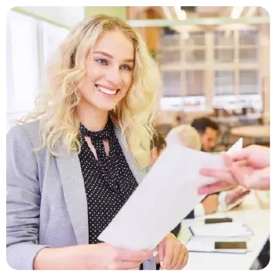 Grinning blonde in business clothes hands a piece of paper to a hand sticking out of the picture.