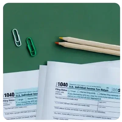 Against a green background are tax documents to be filled out and two pens and paper clips each.