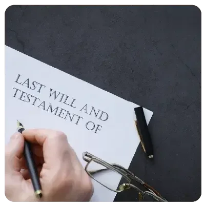 Ein weißer Zettel mit der Aufschrift "Last will and testament of" liegt vor einem dunkelgrauen Hintergrund. Eine Hand hält einen Stift in der Hand, bereit zum Ausfüllen.