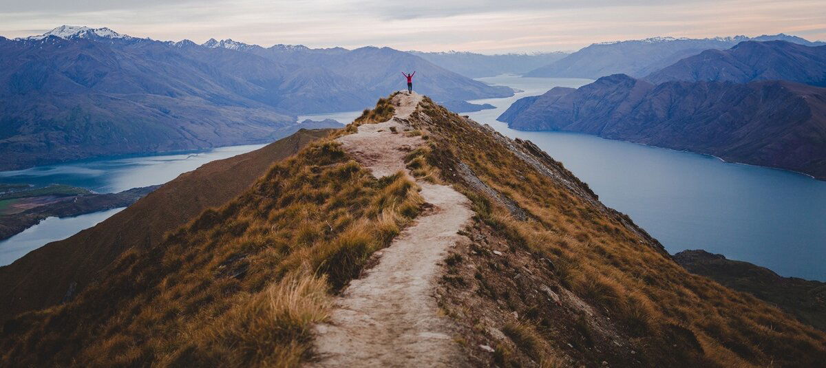 Person standing at the end of a path looking over a sea and mountains