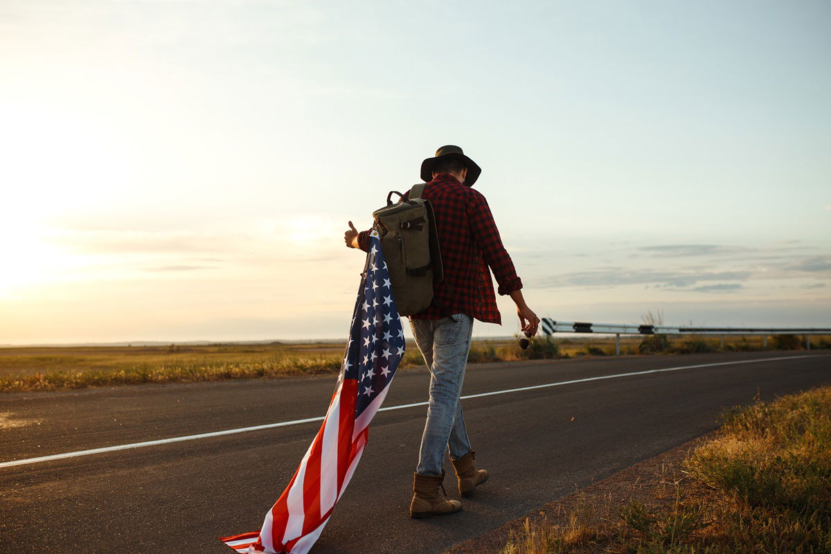 Man walking with backack and US flag alongside highway