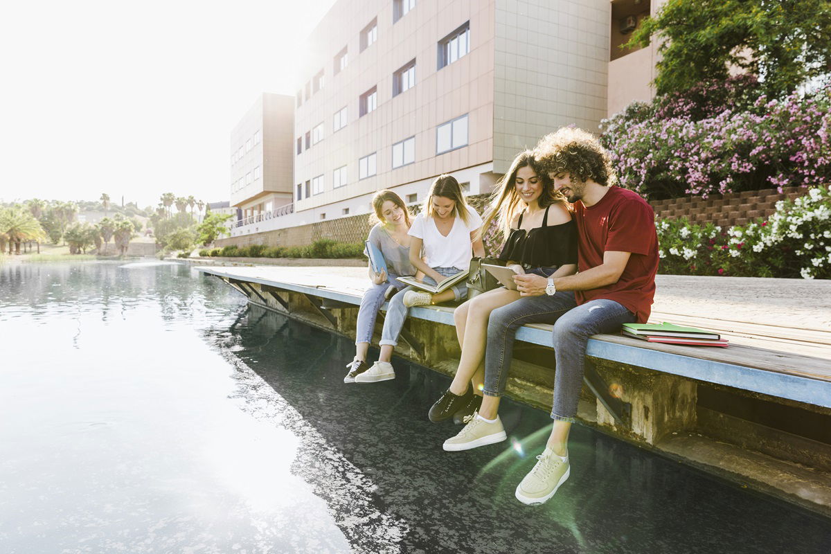 Junge Menschen sitzen an einem Fluss vor einer Universität und lernen.